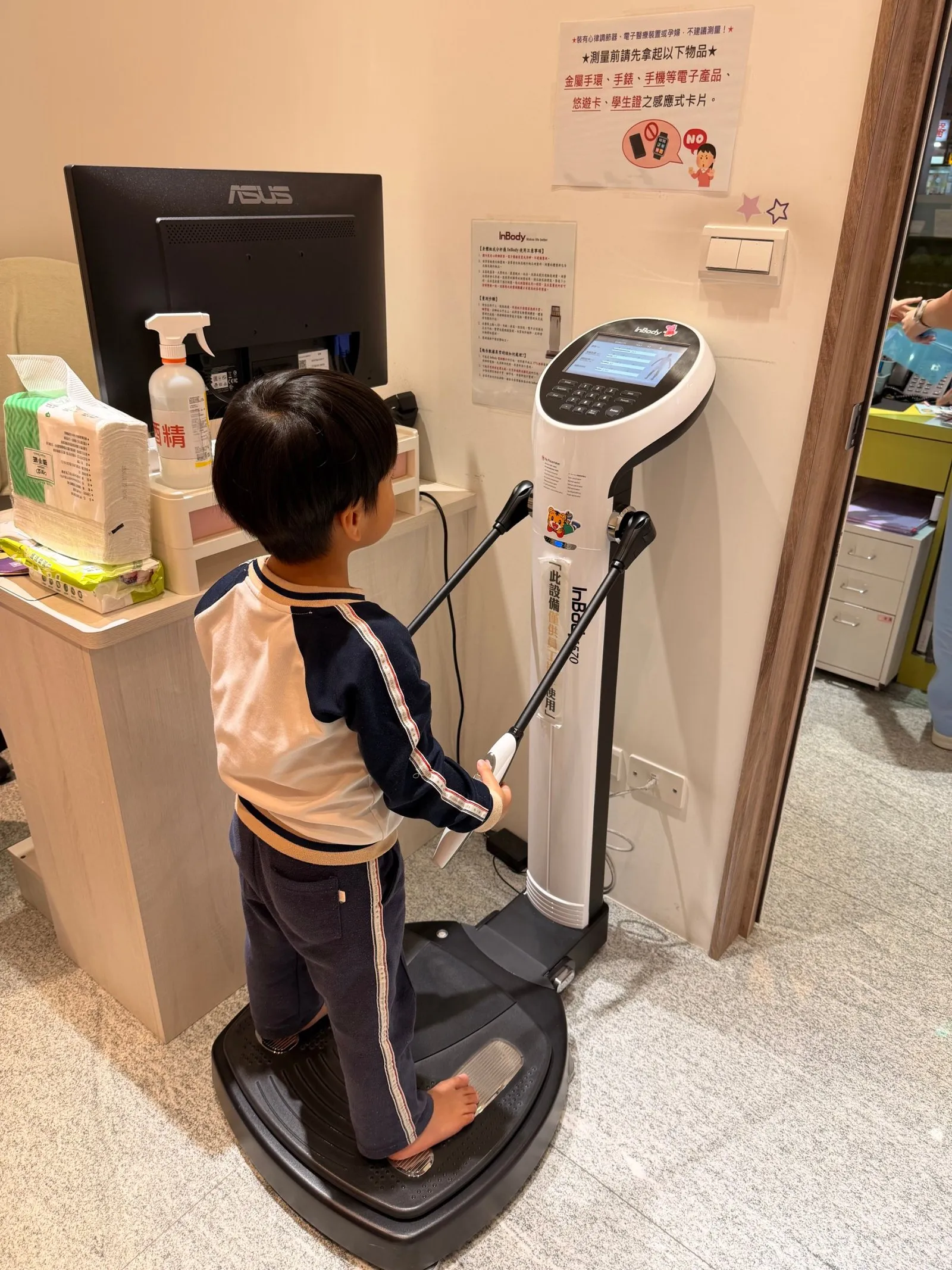 孩子光腳站在 InBody 體組成分析儀上,雙手握著兩側把手,牆上貼著測量前要拿下手錶與感應卡片的提醒
